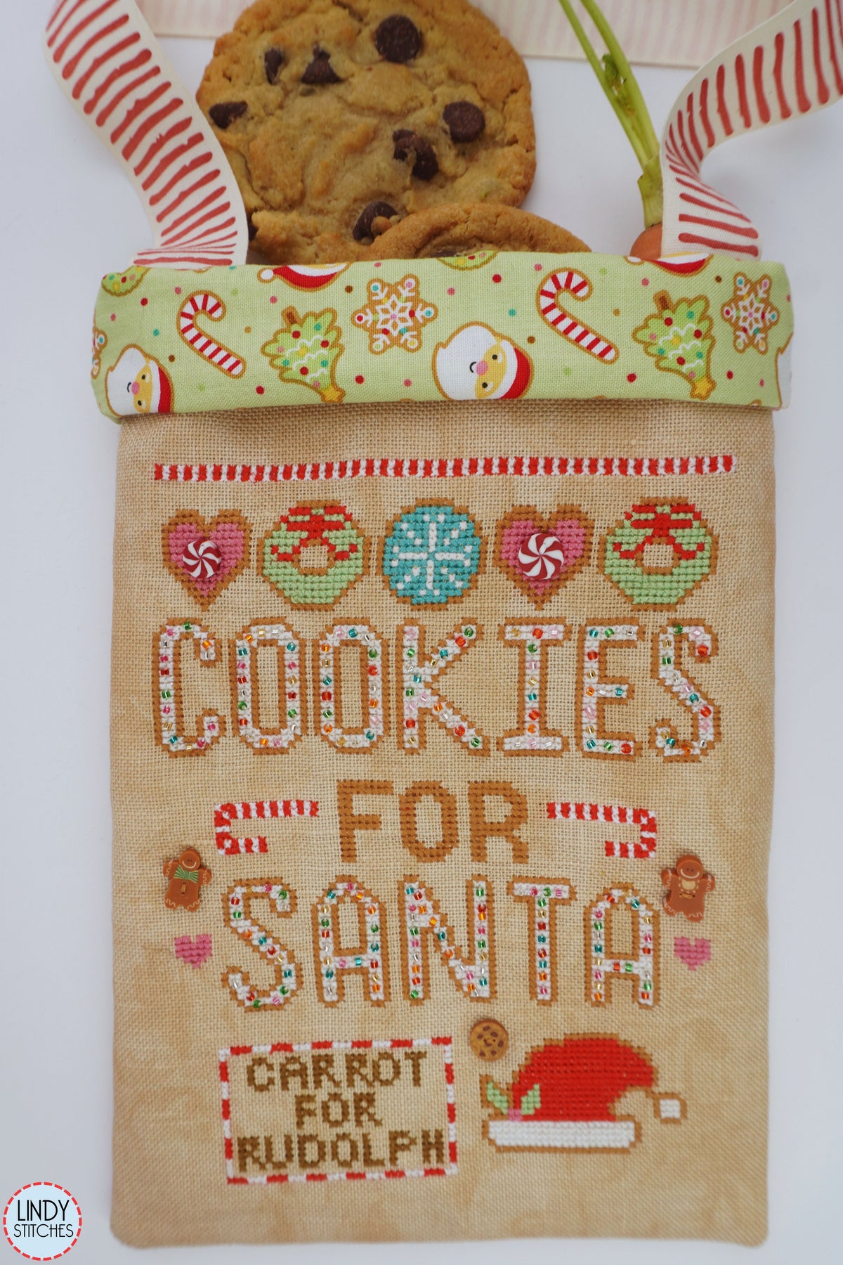 A Christmas-themed burlap cookie bag with embroidered 'COOKIES FOR SANTA' and 'CARROT FOR RUDOLPH' text, decorated with candy canes, snowflakes, and a Santa hat. A chocolate chip cookie and a green stem peek out from the top, which is finished with red-and-white striped ribbon handles.